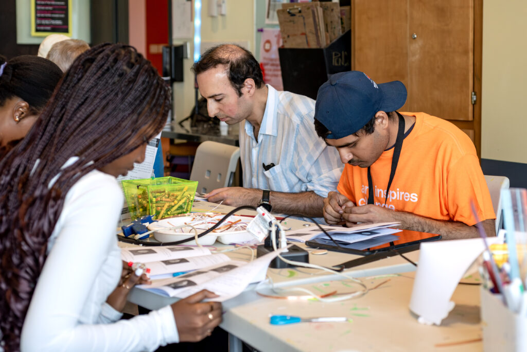 Five people leaned over a table, focused on the projects in front of them. They all have an instruction booklet open and the table is filled with small electronic parts, copper tape, extension chords, crayons, and glue guns